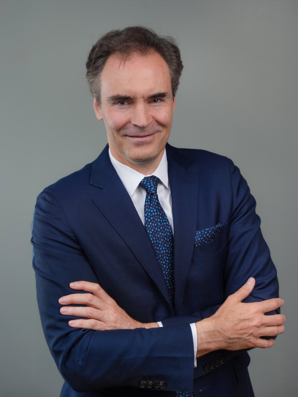 A man in a blue suit with a patterned tie and pocket square stands with his arms crossed, smiling, against a plain gray background.