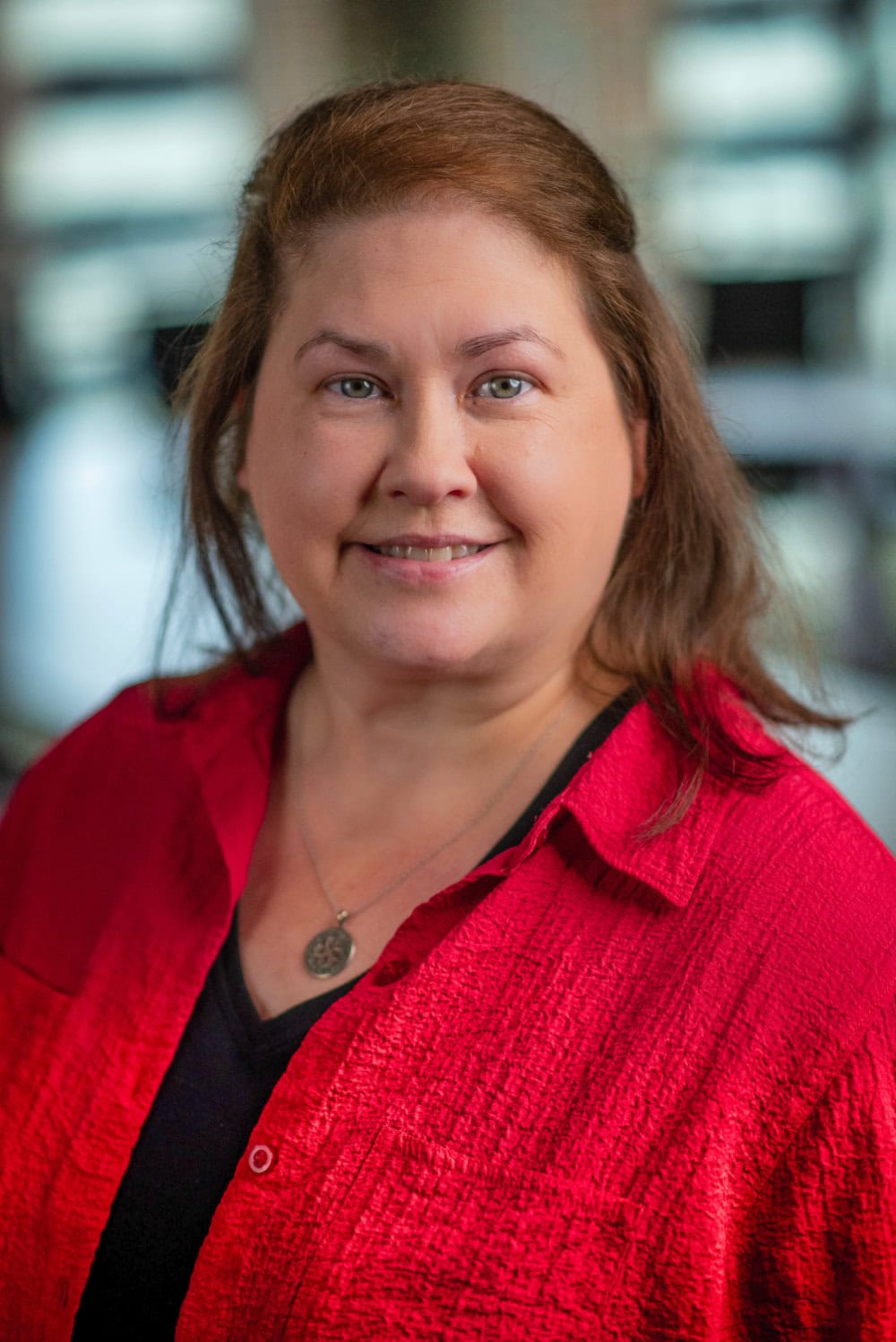 A woman with long brown hair, wearing a red shirt over a black top and a necklace, smiles at the camera with a blurred background.