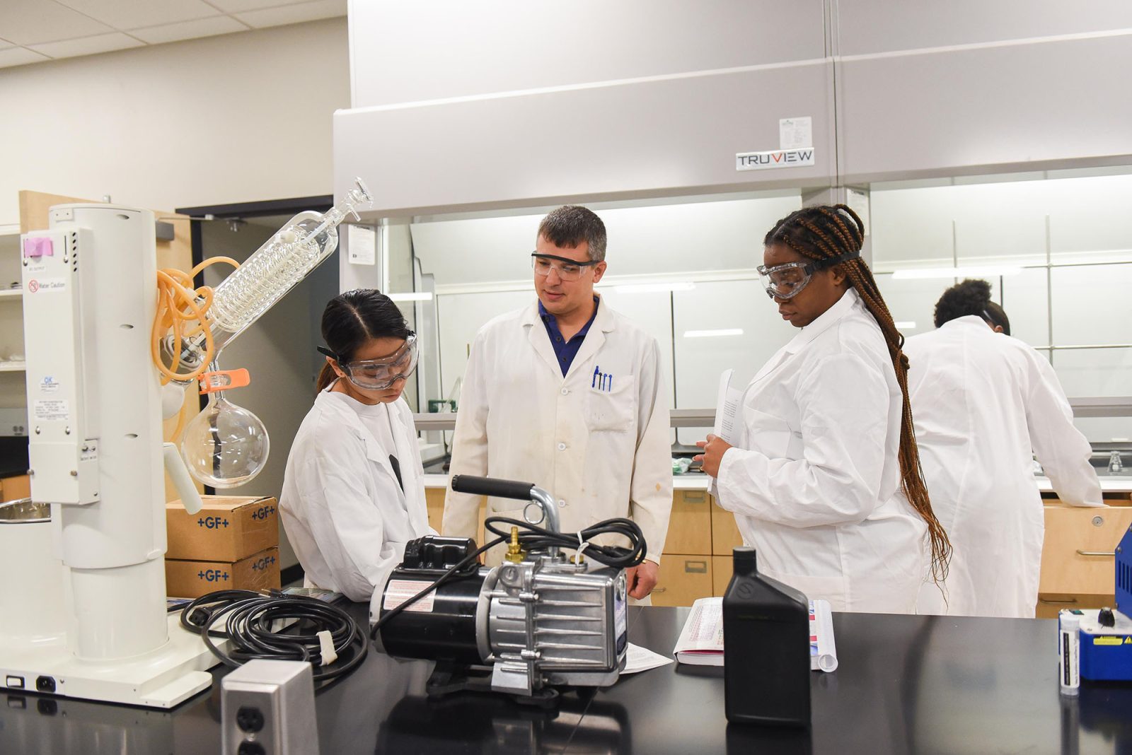 Three people in lab coats and safety goggles stand around a lab bench in a science laboratory, discussing documents and equipment.