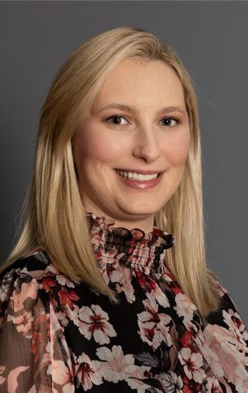A woman with straight, shoulder-length blonde hair smiles at the camera. She is wearing a black blouse with pink and white floral patterns and a ruffled collar, against a gray background.