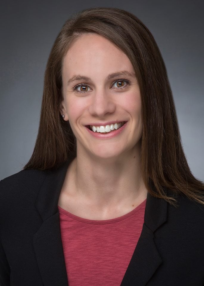 A woman with straight brown hair wearing a black blazer and a red top smiles at the camera against a gray background.