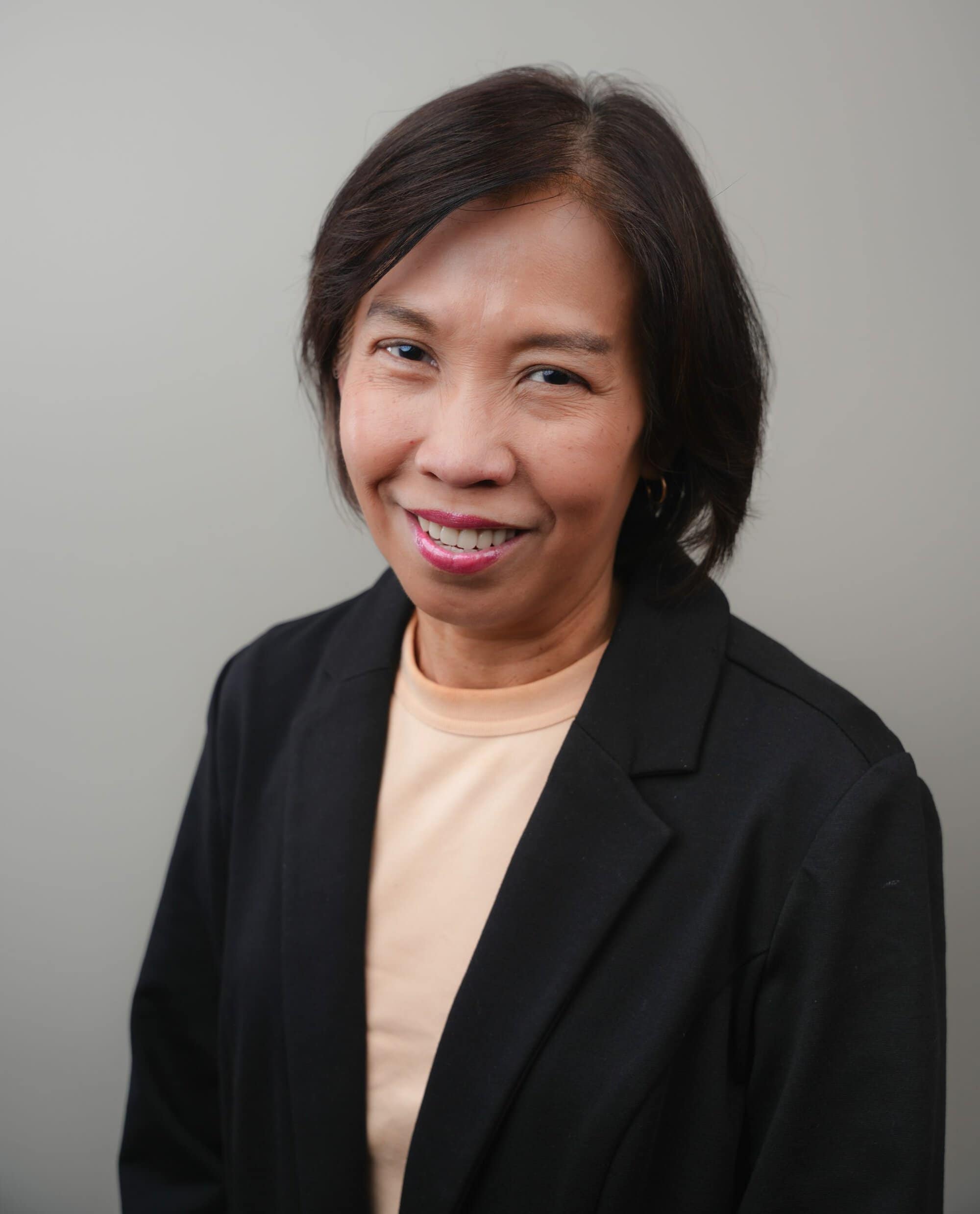 A woman with short dark hair wearing a black blazer and peach top stands in front of a plain gray background, smiling slightly.