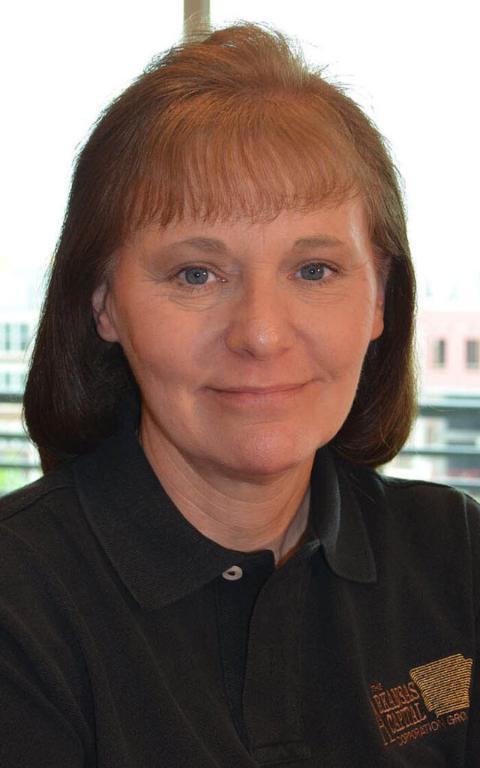 Middle-aged woman with brown hair and blue eyes wearing a black collared shirt, sitting indoors with a window and buildings in the background.