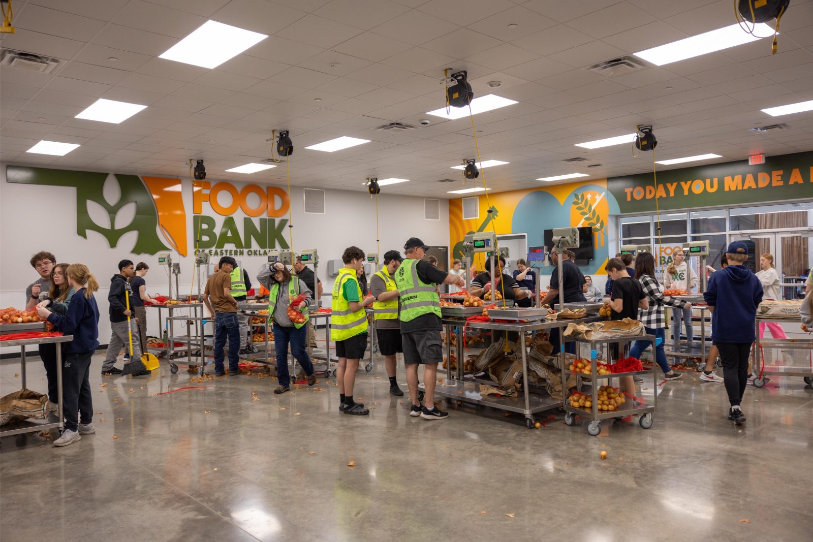 Volunteers in high-visibility vests sort and pack food items at a food bank warehouse with tables and produce bins.