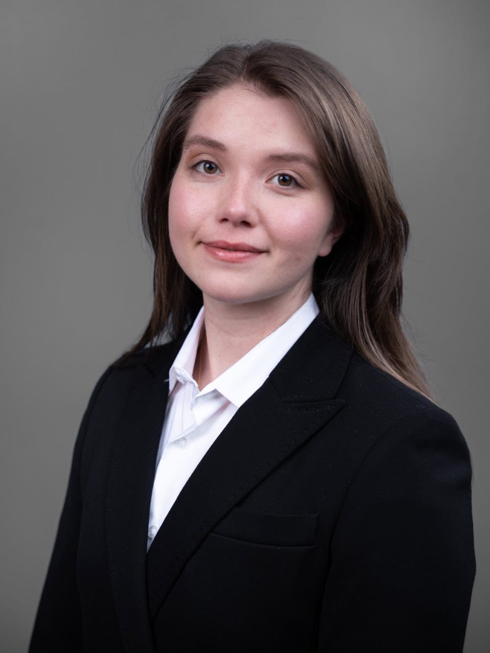 A woman with long brown hair wearing a white shirt and black blazer poses for a professional headshot against a plain gray background.