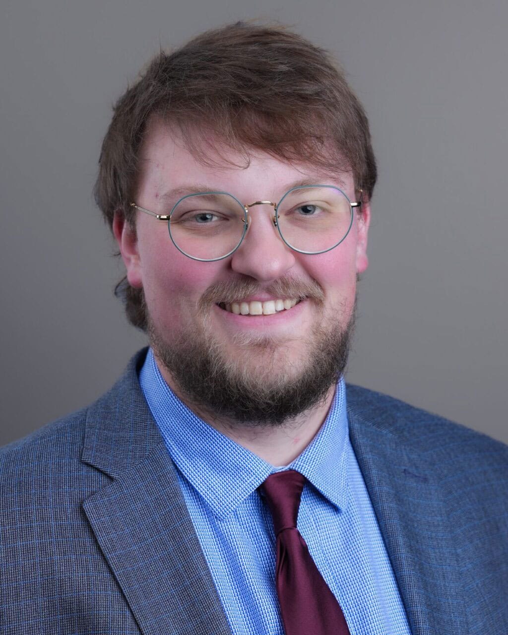 Man with light brown hair, beard, and glasses wearing a blue suit, light blue shirt, and burgundy tie, smiling at the camera with a neutral background.