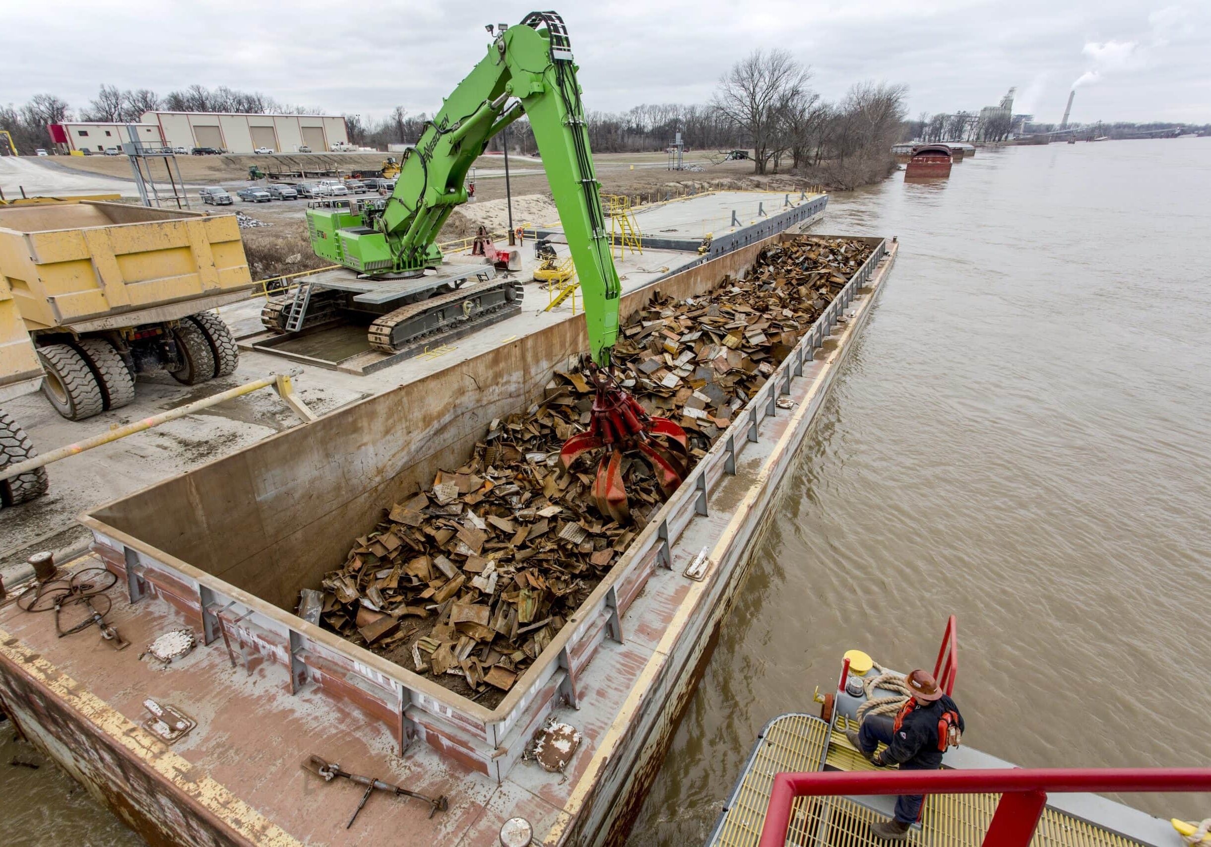 A green excavator loads scrap metal onto a barge docked on a river, while a worker stands nearby on a metal platform.