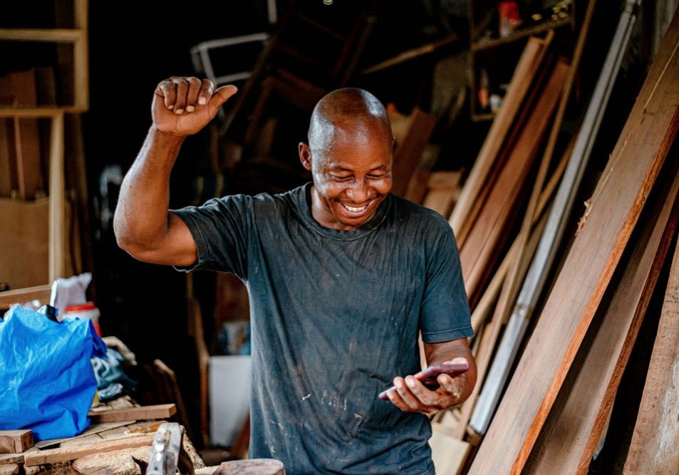 A man in a workshop smiles and raises his arm while looking at a smartphone, surrounded by wooden boards and tools.