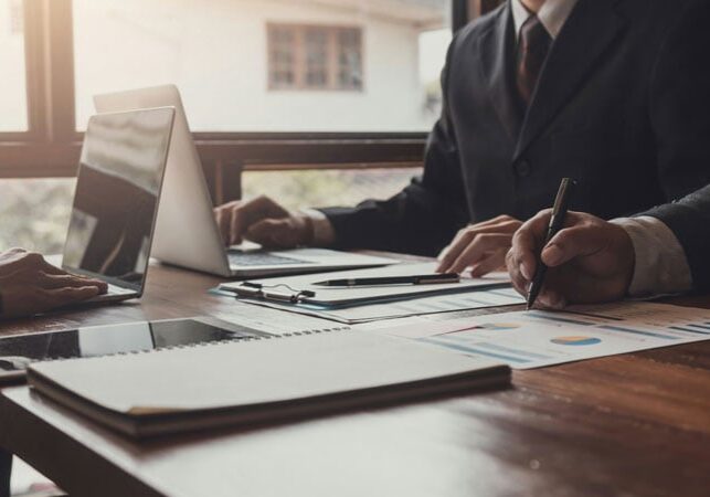 Two people in business attire work at a desk with laptops, documents, and charts, reviewing papers and taking notes in a sunlit office.