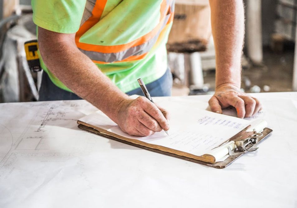 A person wearing a high-visibility shirt writes on a clipboard with papers, standing over a table with construction plans.