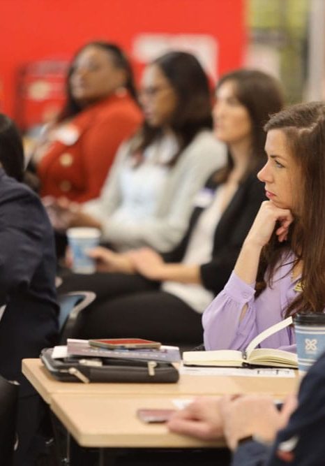 A group of adults sit at tables, attentively listening to a presentation, with notebooks, coffee cups, and folders in front of them.