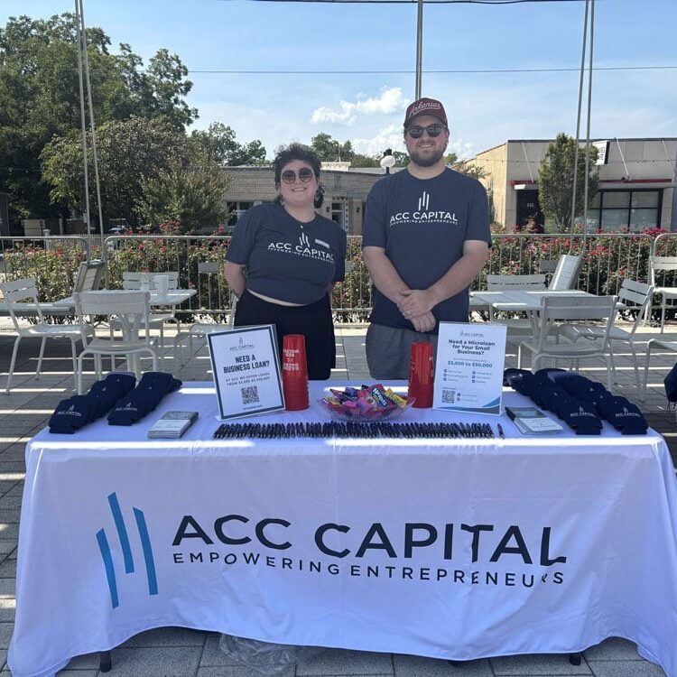 Two people stand behind an ACC Capital booth with promotional items and informational signs on a table, outdoors on a sunny day.