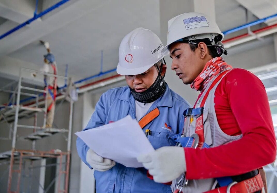 Two construction workers wearing safety gear review documents at a construction site with scaffolding and exposed piping in the background.