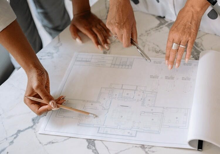 Two people review and point at a building floor plan on a marble table, holding a pencil and a pen.