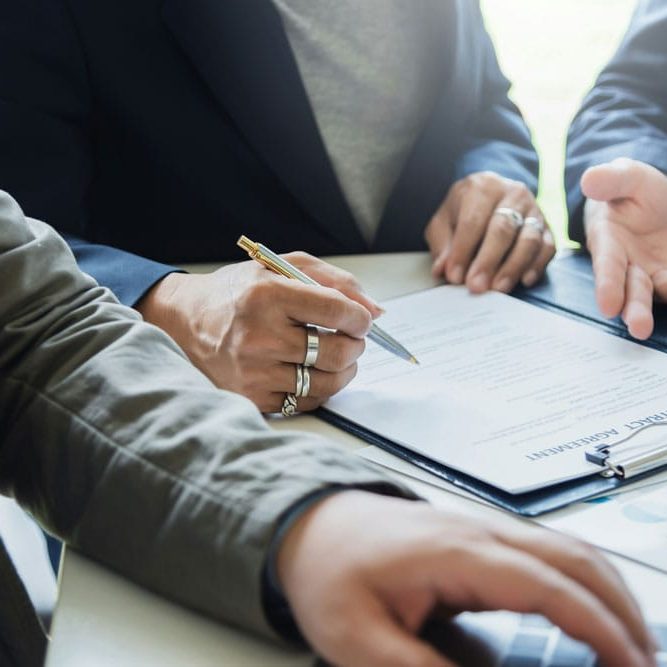 Two people in business attire review and sign a contract agreement at a desk with charts and documents.