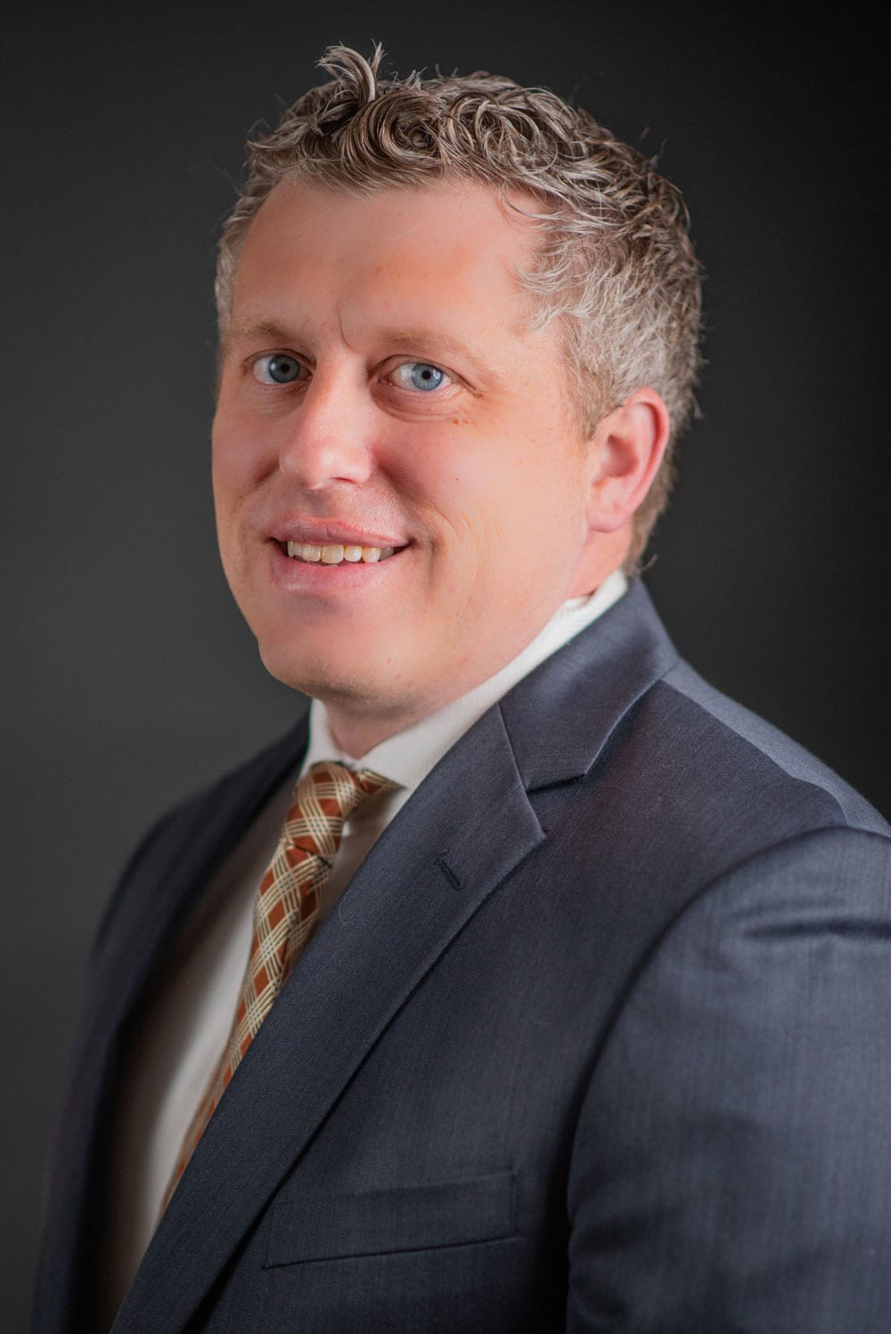 Man in a gray suit and patterned tie poses against a dark background, facing slightly to the side and smiling.