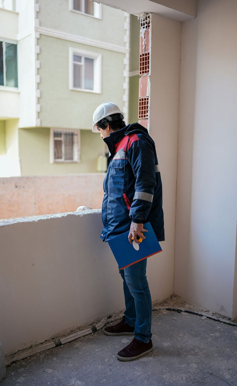 A person wearing a hard hat and holding blue folders stands on a balcony of a building under construction, looking down.