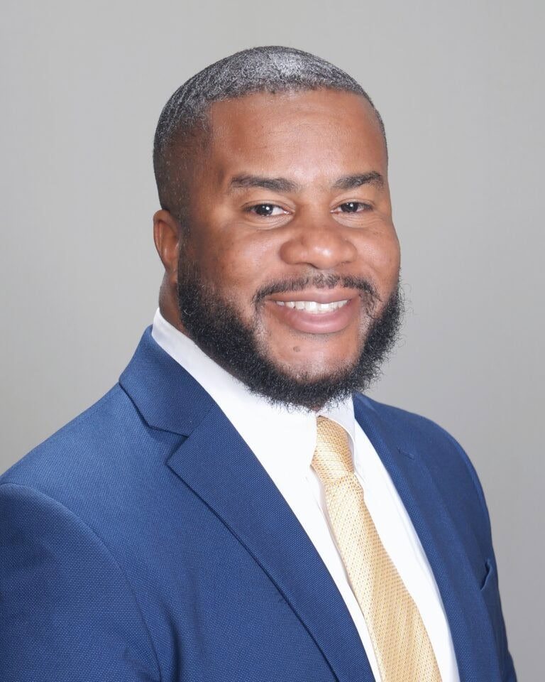 A man wearing a blue suit, white shirt, and yellow tie smiles at the camera against a plain light gray background.