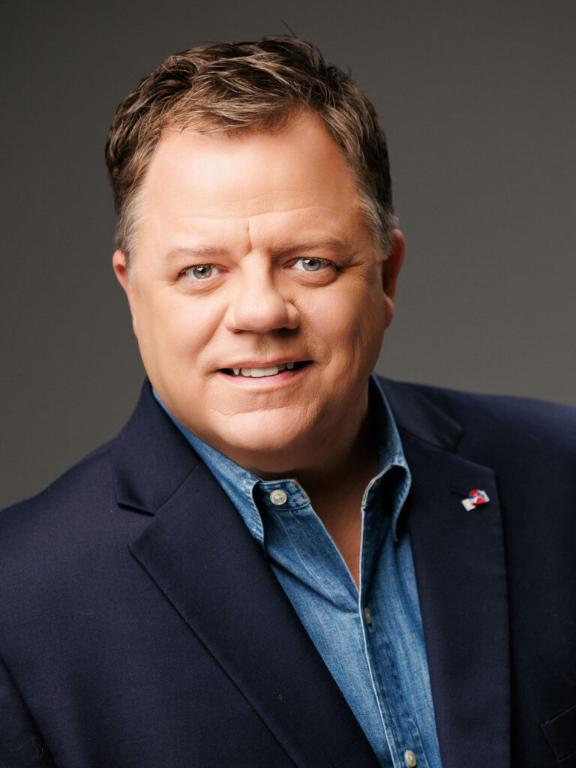 A man with short brown hair is wearing a navy blazer over a blue collared shirt, smiling at the camera against a plain background.