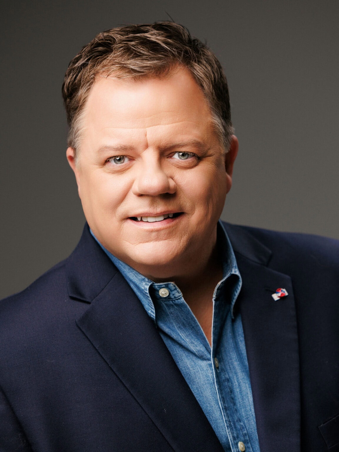 A middle-aged man with short brown hair wearing a navy blazer over a blue shirt, smiling at the camera against a plain background.