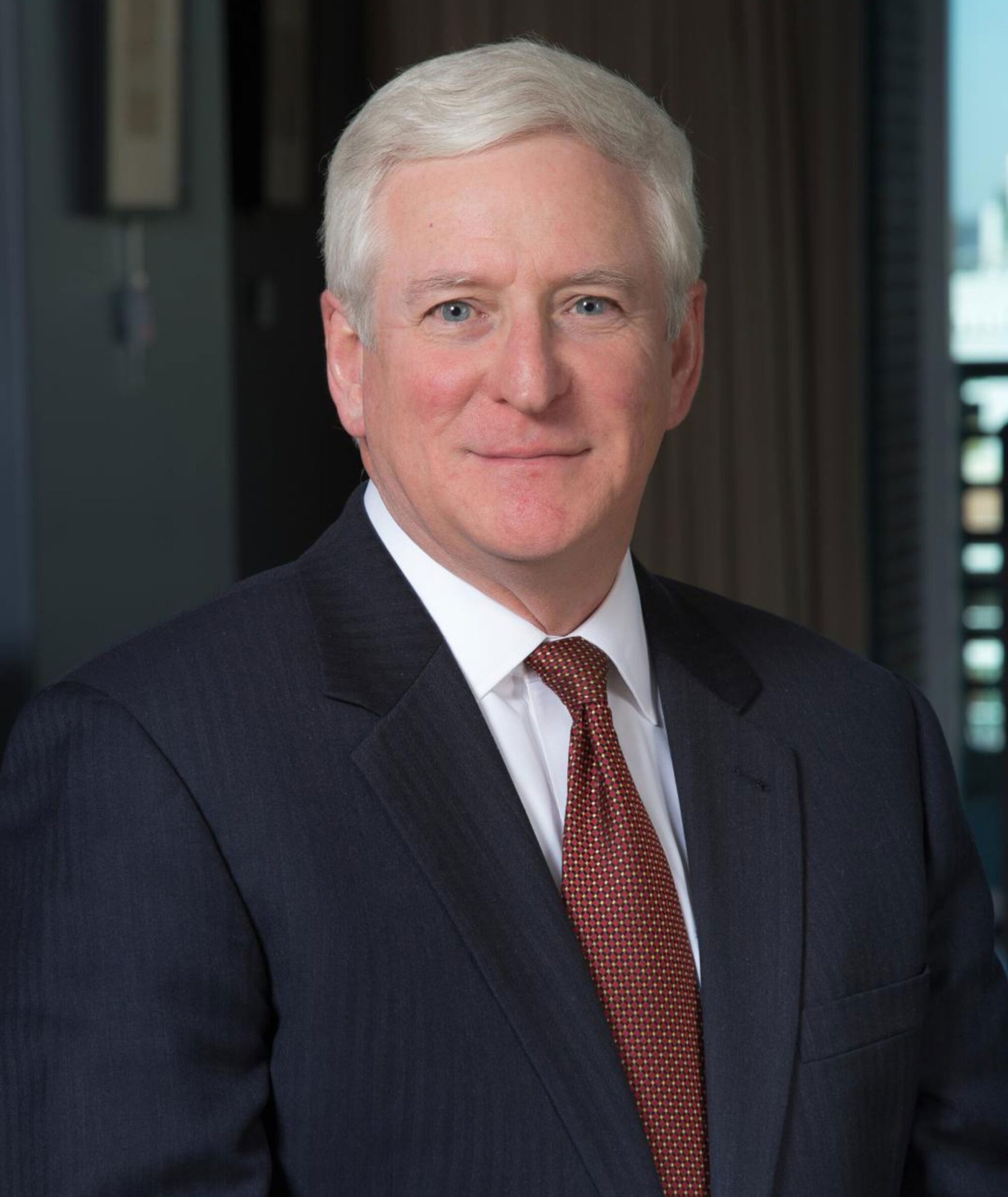 A middle-aged man with short white hair wearing a dark suit, white shirt, and red patterned tie poses for a professional portrait indoors.
