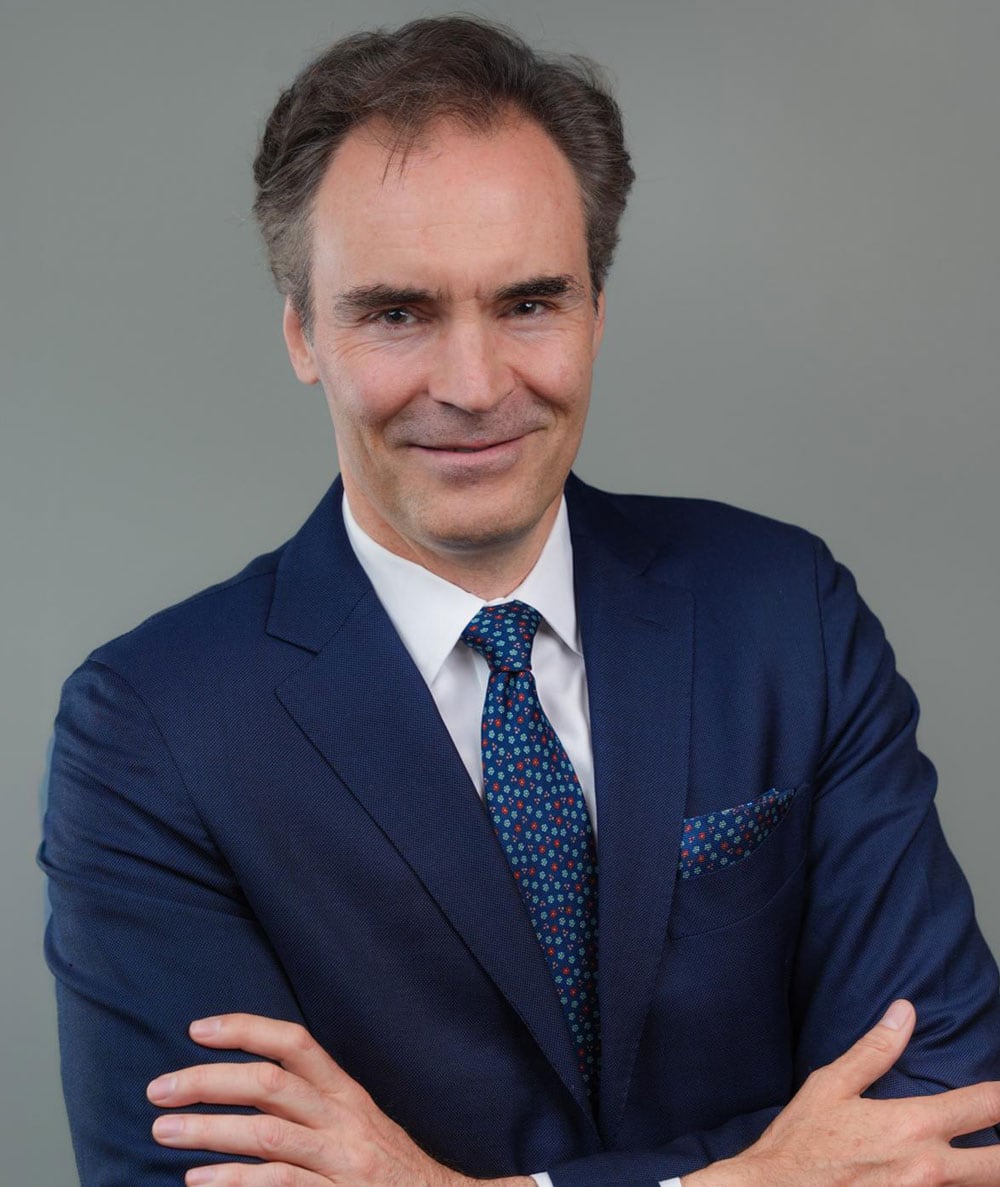 Man in a blue suit with a patterned tie and pocket square, standing against a plain gray background with arms crossed, smiling at the camera.